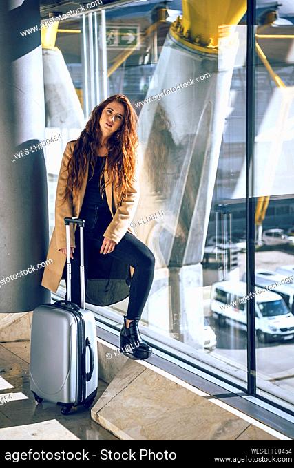 Confident businesswoman standing with luggage by window at airport departure area