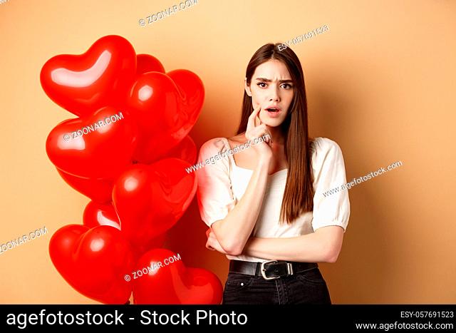 Valentines day and love concept. Confused and shocked girl staring displeased at camera, standing near romantic red hearts balloons on beige background
