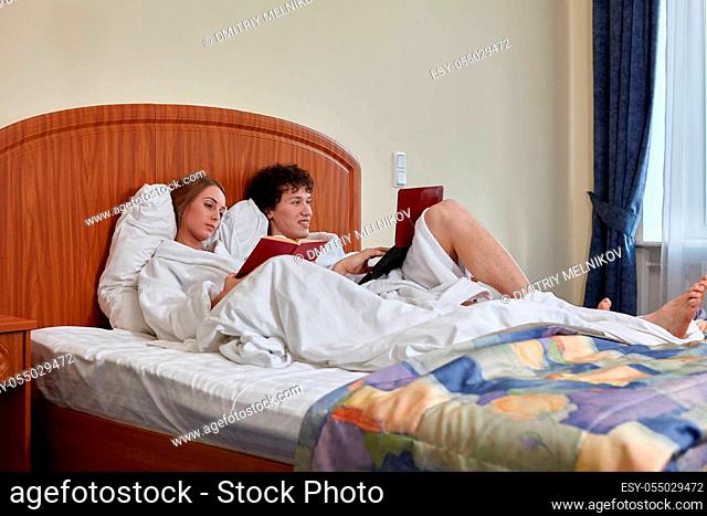 Young family with laptop and book are lying on bed in room at home. Weekend or isolation period, social distancing. Remote learning and work