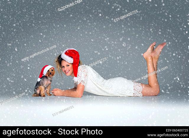 Beautiful happy girl in lace dress and red santa cap lying with pretty yourkshire terrier in christmas hat. Studio shot over grey background with falling snow