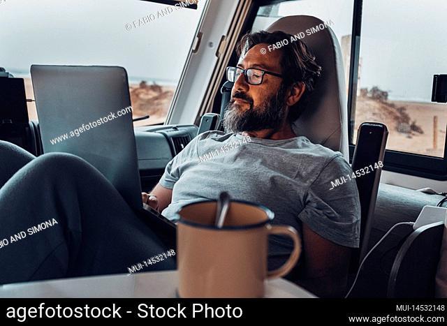 Digital nomad new modern job lifestyle with handsome adult man working and relaxing inside a camper van with beach and nature outside