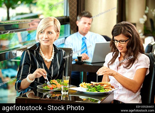 Young women sitting at table, eating sandwich and salad in restaurant, smiling