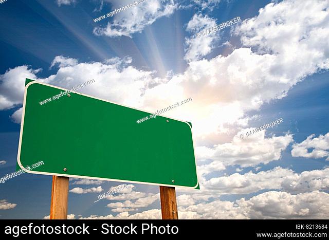 Blank green road sign over dramatic blue sky with clouds and sunburst, ready for your own message and room for copy on clouds