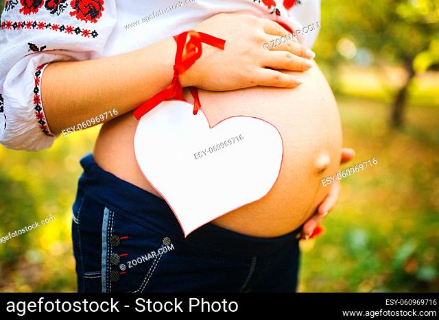 a pregnant woman with her husband embrace the belly, dressed in a traditional Ukrainian outfit and hold a paper heart with space for text