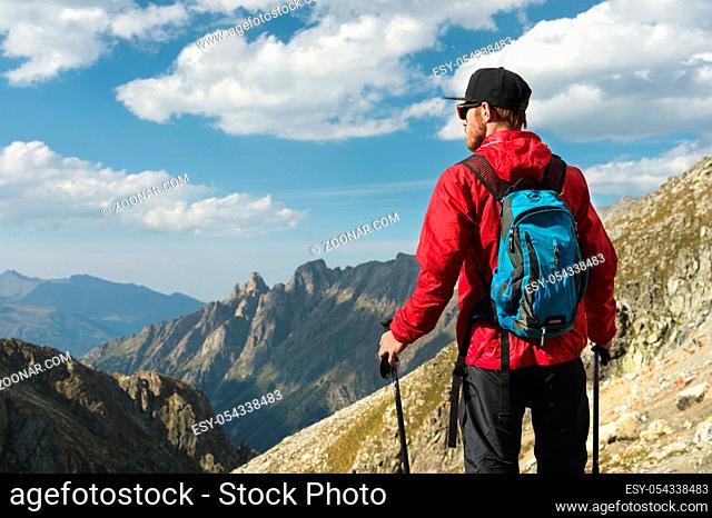 A bearded man in sunglasses and a cap with a backpack stands on top of a rock and looks into a rocky valley high in the mountains