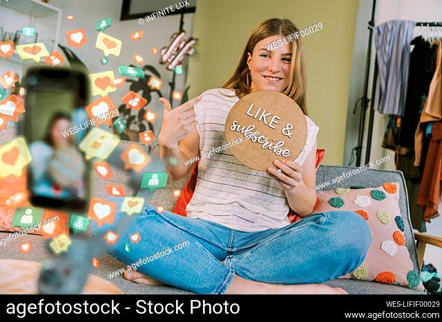 Female influencer showing like and subscribe cardboard while vlogging through mobile phone at home