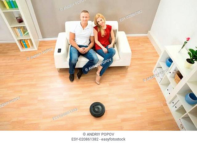Couple On Sofa With Robotic Vacuum Cleaner On Floor