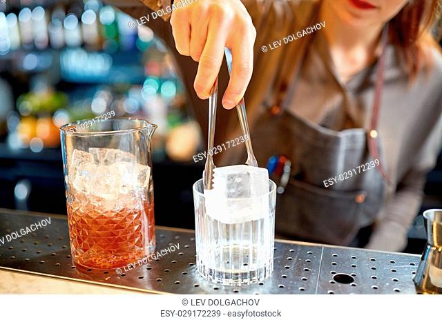 alcohol drinks, people and luxury concept - woman bartender with tongs adding ice cube into glass and preparing cocktail at bar counter
