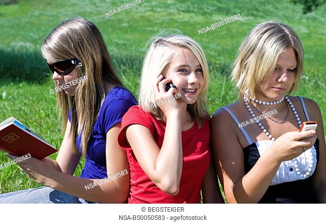 three teenage girls sitting with mobile phones and book in meadow