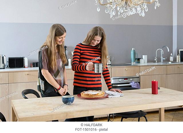 Young woman watching sister sprinkling tart with icing sugar in kitchen