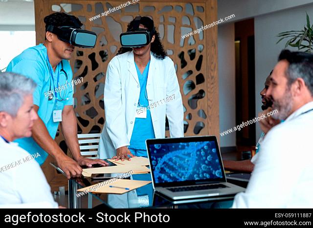 Diverse male and female doctors wearing face masks sitting at table and using vr glasses