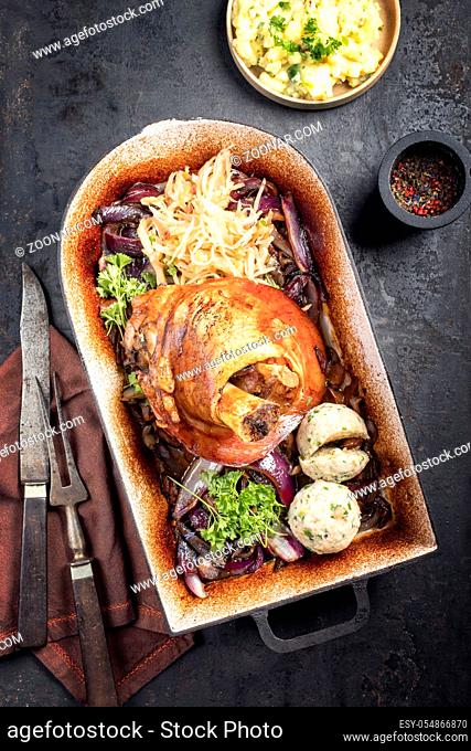 Traditional Bavarian schweinshaxe with onion, potato salad and sauerkraut as top view in a rustic stewpot on an old board