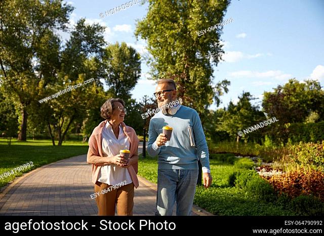 Happy elderly family having coffee break walking in park. Romantic aged mature man and woman looking happy enjoying healthy lifestyle on pension