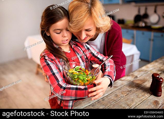 Little girl holding salad bowl in kitchen. Granny helping her
