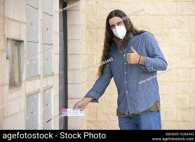 Caucasian male with long hair and full beard wearing a mask and putting official election mail in ballot in the outgoing mail slot at his apartment building
