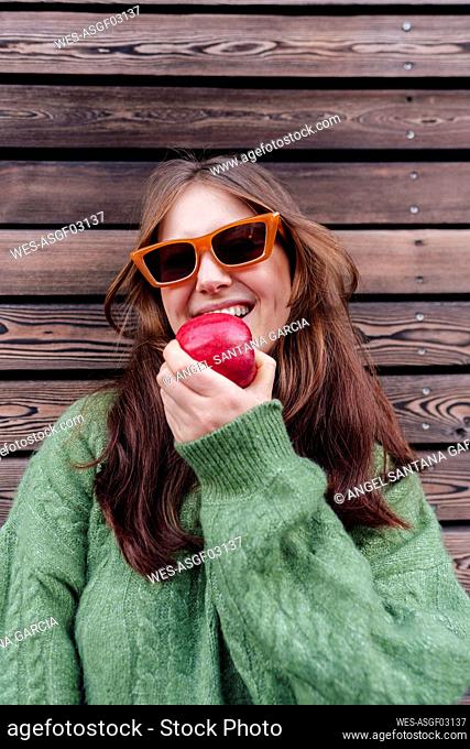 Happy woman wearing sunglasses eating apple in front of wooden wall