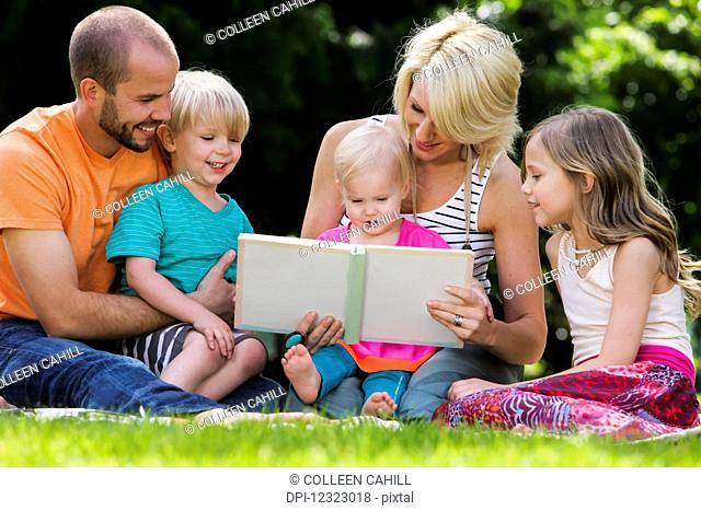 A family with three children sit on the grass reading a book; Oregon, United States of America