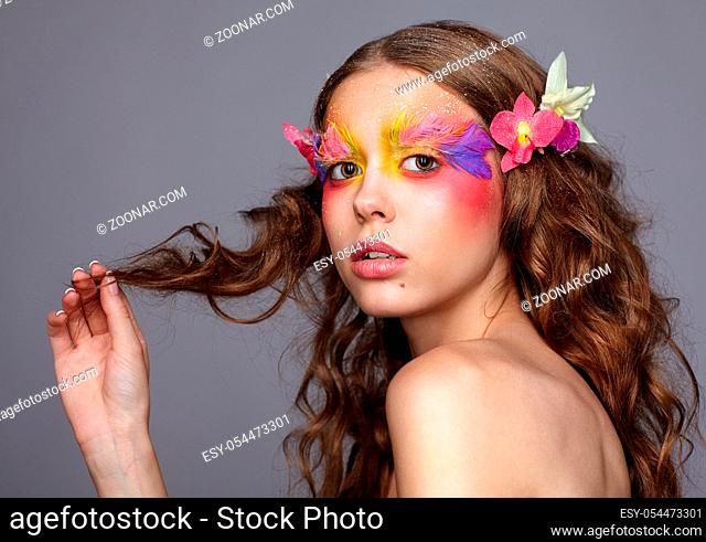 Portrait of teen girl with hand near face. Young female with unusual stylish make-up and false fashion feather eyelashes. Orchid flower in wavy hair