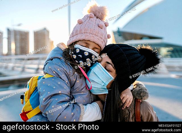 Mother and daughter wearing knit hat and face mask hugging while standing outdoors