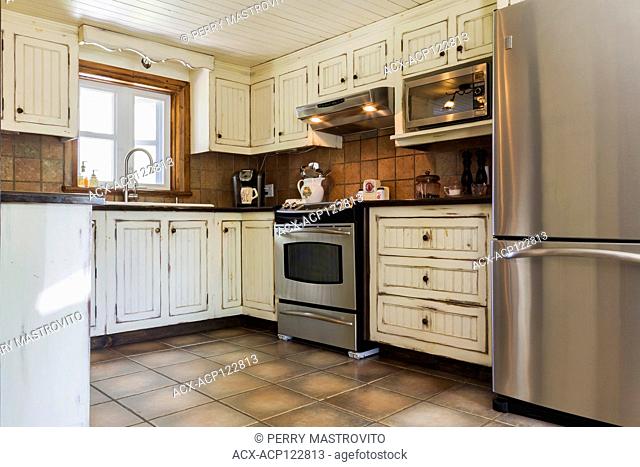 Kitchen with cream coloured antique finish wooden cabinets, ceramic countertops and tile floorinside an old 1892 Canadiana cottage style home, Quebec, Canada
