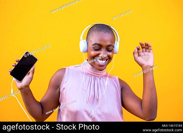 happy african or american woman dancing and vibing alone with her phone in her hand and with headphones on her head listening music having fun -close up and...