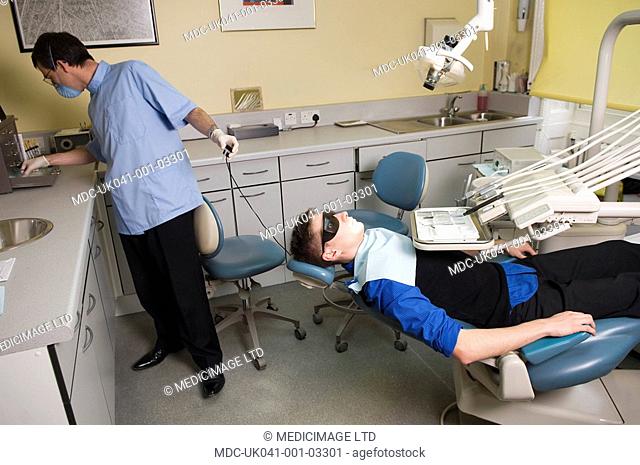 Dentist wearing safety glasses and mouth mask prepares to xray the teeth of young male patient, also wearing special protection glasses