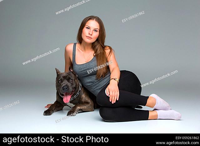 Beautiful sporty young woman sitting on floor and hugging adult grey amstafford terrier dog. Studio shot over gray background. Copy space
