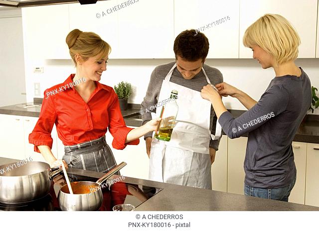 Two young women helping a young man in wearing an apron in the kitchen