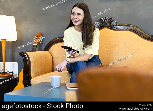Happy young woman holding smart phone while sitting on sofa in modern cafe