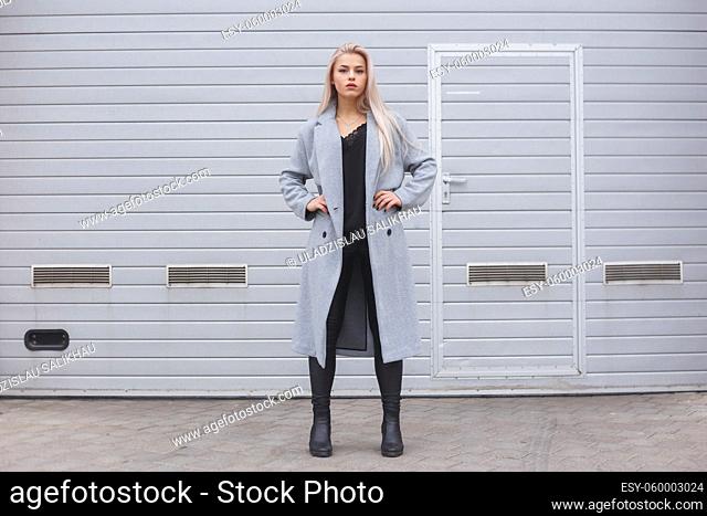 Elegant young woman wearing gray coat posing against rough street wall, minimalist urban clothing style
