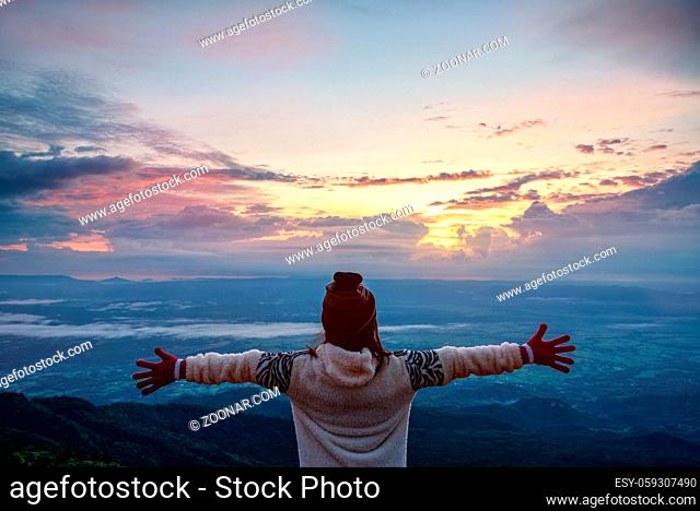Woman tourist in a sweater dress standing outstretched arms, happy while watching the beautiful nature landscape of the forest and mountain during the sunrise...