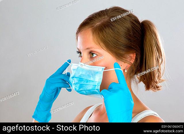 Young woman with blond hair putting on face mask from side view. Portrait of female wearing blue rubber gloves and attaching protective respiratory filter...