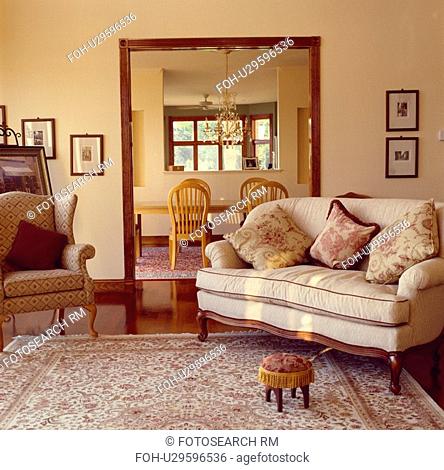 Patterned armchair and cream sofa with toile-de-Jouy cushions in front of doorway in livingroom with small footstool on patterned carpet