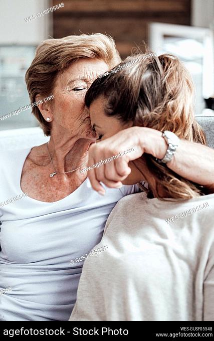 Caring grandmother kissing granddaughter on forehead while sitting at home