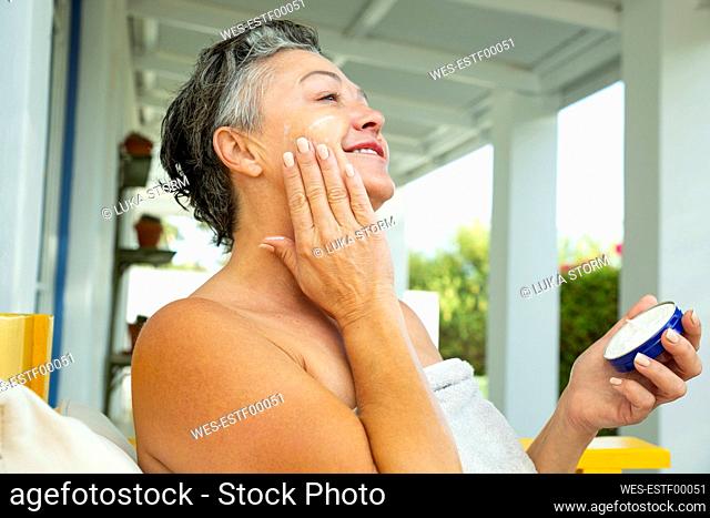 Smiling mature woman applying moisturizer on face sitting at home