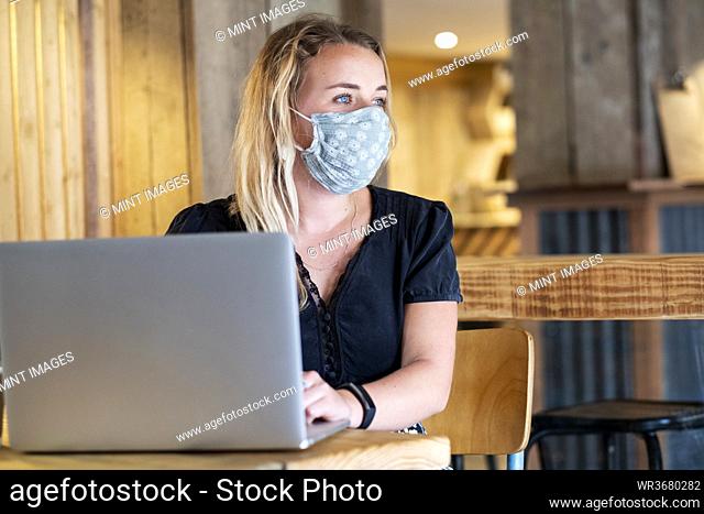 Young blond woman wearing blue face mask, sitting at table, using laptop computer