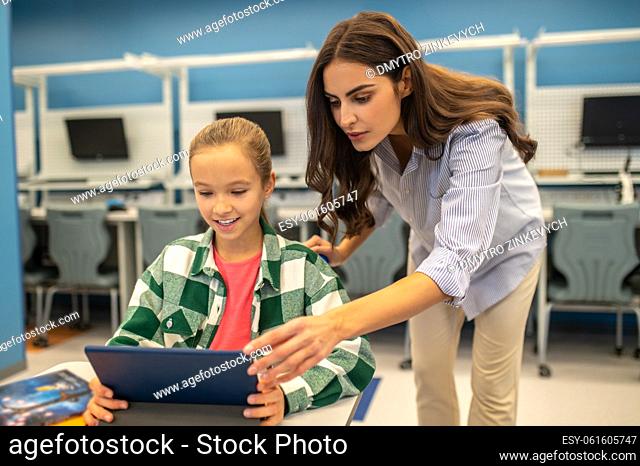 Learning process. Attentive involved woman teacher leaning over touching tablet of smiling girl sitting at desk in classroom