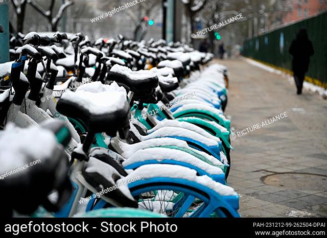 12 December 2023, China, Peking: Snow-covered rental bikes from several providers are parked on a sidewalk. Many people in Beijing use the rental bikes to get...