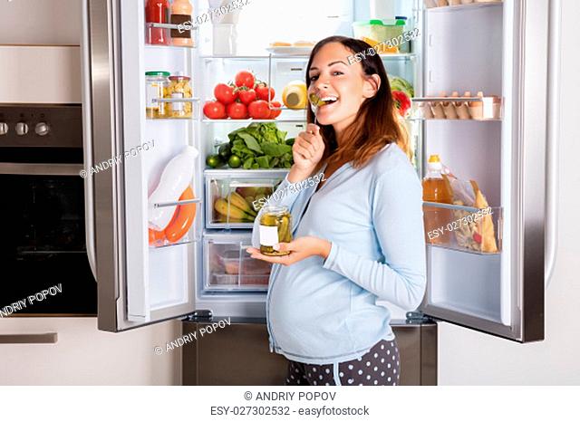 Young Pregnant Woman Enjoy Eating Jar Of Pickle In Front Of Open Refrigerator In Kitchen