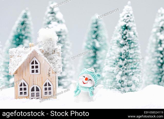 Small decorative snowman with gift near wooden house in fir forest under falling snow