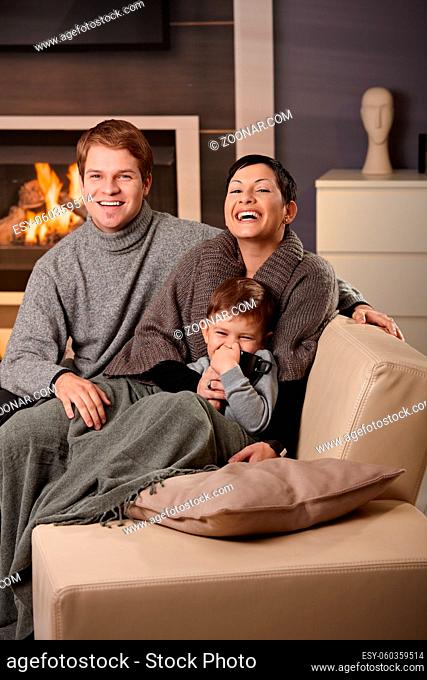 Happy family sitting on couch at home in front of fireplace, looking at camera, laughing