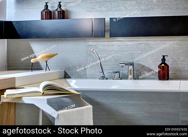 Gray tiled bathroom with black shelves on the wall. There is a bath with chrome shower and faucet, pump bottles, stand with a towel and a book