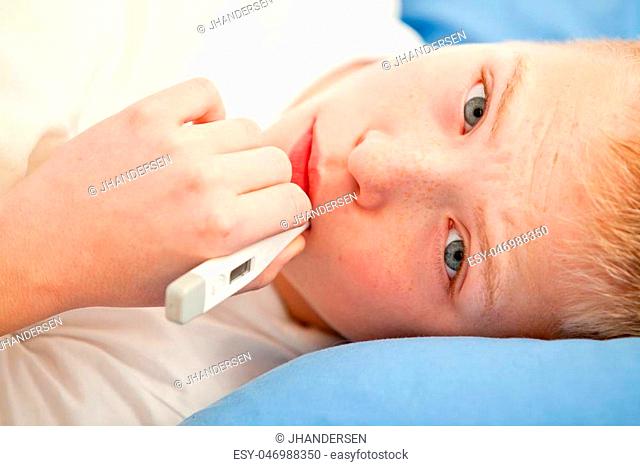 Close up view on sick boy using white digital thermometer while laying down on blue linen pillow