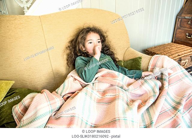 A six year old girl holding a toy rabbit sitting on a sofa wrapped in a blanket