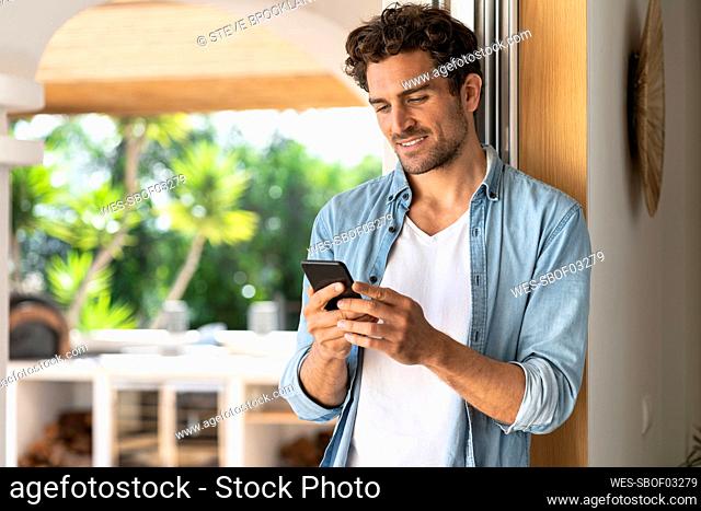 Smiling man looking at smart phone while standing at doorframe