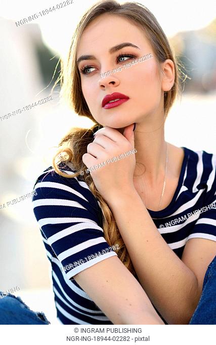 Blonde woman, model of fashion, sitting on a bench in urban background. Beautiful young girl wearing striped t-shirt and blue jeans in the street