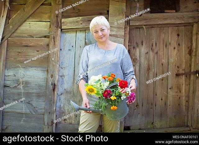 Smiling mature woman holding bouquet of multi colored flowers in galvanized water can