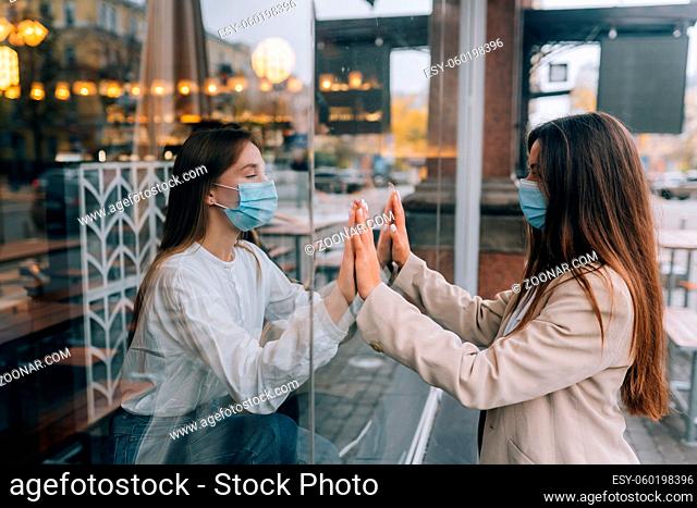 Two young girls in protective masks opposite each other, a window between them. The concept of self-isolation in the context of Epidemai