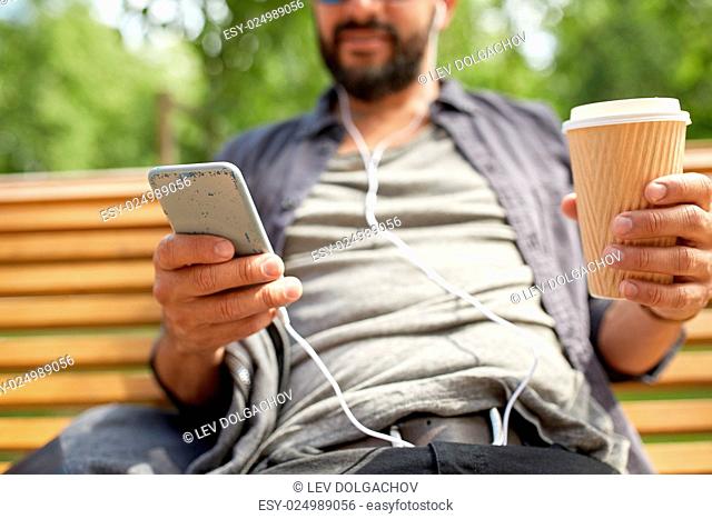 people, drinks, technology, leisure and lifestyle - close up of man with earphones and smartphone drinking coffee from disposable paper cup on city street bench