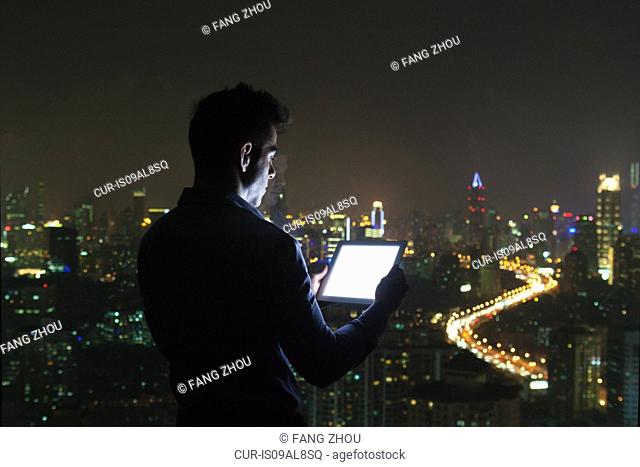 Silhouetted young businessman looking at digital tablet in front of skyscraper office window at night, Shanghai, China